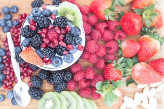 Healthy nutritious snack made of a bowl of raspberries, blackberries, blueberries, pomegranate, strawberries and kiwi, surrounded by fresh fruits on a wooden table