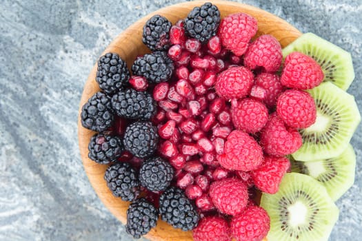 Healthy breakfast of fresh fruit with sliced tropical kiwifruit arranged with whole blackberries and raspberries and cleaned pomegranate seeds in a wooden bowl, overhead view on a grey stone counter