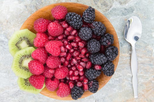 Delicious nutritional snack made of fresh slices of kiwi, seeds of pomegranate, blackberries and raspberries, in a wooden bowl, next to a metallic spoon, on a table, high-angle shot