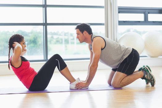 Full length side view of a male trainer helping young woman do abdominal crunches in the bright gym