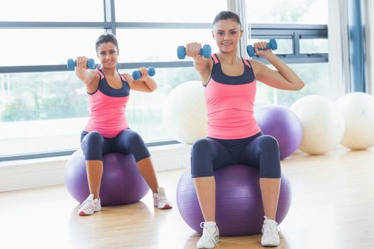 Two fit young women exercising with dumbbells on fitness balls in the bright gym