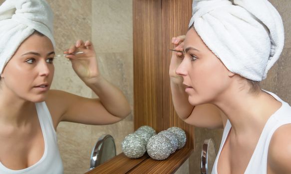 Beautiful young woman plucking her eyebrows with tweezers in front of mirror on the bathroom