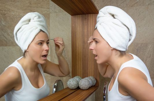 Beautiful young woman suffering pain when plucking eyebrows with tweezers in front of the mirror