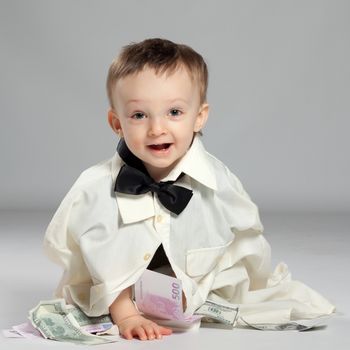 Boy toddler businessman sitting dressed in grown-shirt with bow tie, in front of him has a pile of money and enjoys