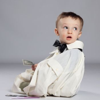 Boy toddler businessman, sitting next to a pile of money dressed in grown-shirt with bow tie, holding in hand the dollars