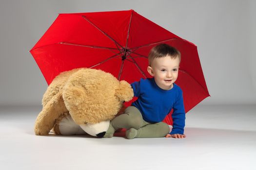 Toddler boy sitting under a red umbrella next to a large teddy bear