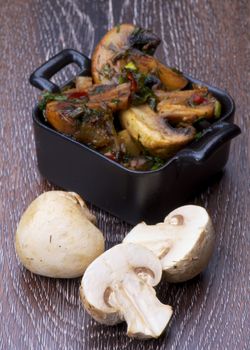 Delicious Roasted Edible Champignon Mushrooms in Black Cast-iron Stew Pots and Halves of Raw Mushrooms isolated on Wooden background