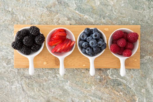 Assorted fresh berries in taster dishes viewed from above lined up on a wooden board on a stone kitchen counter with blackberries, sliced strawberry, blueberries and raspberries