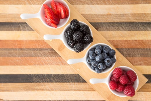 Dishes of fresh berries on decorative striped wood arranged in a diagonal row on a small wooden board including blackberries, raspberries, strawberries and blueberries for a tasty nutritious snack
