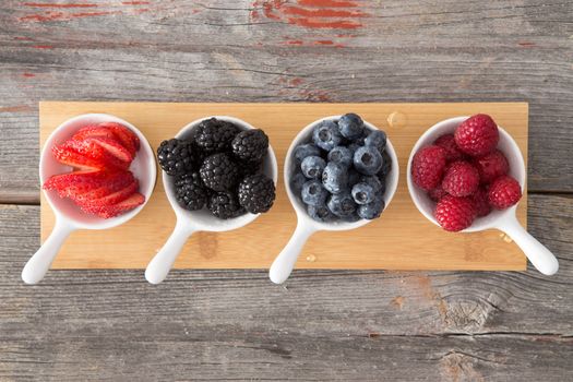 Taster dishes of assorted autumn berries viewed from above arranged on a wooden board on a rustic, kitchen table including whole fresh blueberries, blackberries, raspberries and sliced strawberries