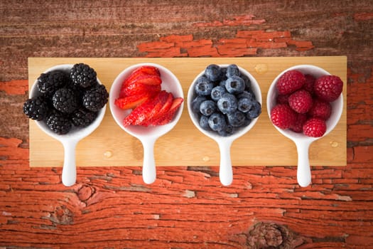 Fresh assorted berries on a grungy wooden counter displayed in small ceramic ramekins including blackberries, blueberries, strawberries and raspberries for a healthy snack or appetizer