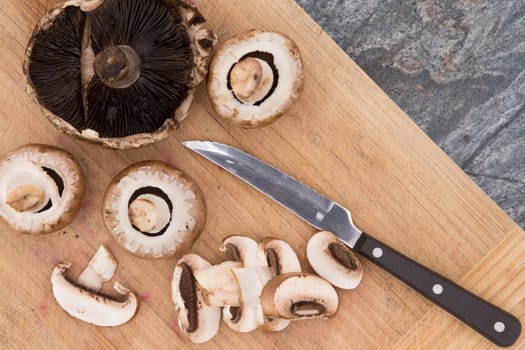 Slicing fresh cultivated agaricus mushrooms for cooking on a wooden chopping board on a stone kitchen counter with a small knife, overhead view