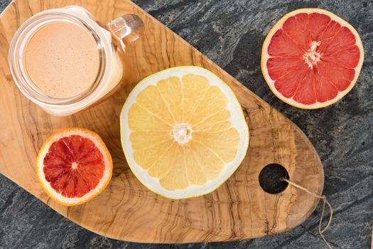 Freshly prepared grapefruit juice with halved ruby and ordinary grapefruit used as ingredients on an old olivewood cutting board with decorative grain , view from above on a stone counter