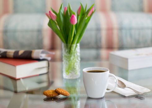 Modern white porcelain cup of black coffee on glass table with spoon and ginger biscuits. Book newspaper and vase of tulips in the background out of focus