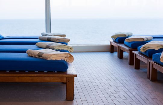 Row of teak reclining couches or benches with cushions and towel by window looking out to sea with horizon in the distance