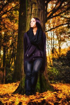 Beautiful young woman with purple hair standing outdoors with her back against a tree and looking up