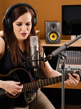 Photo of a beautiful brunette in a recording studio playing an acoustic guitar and singing into a large diaphragm microphone.