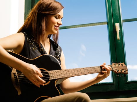 Photo of a smiling woman in her late twenties playing an acoustic guitar by a large window.