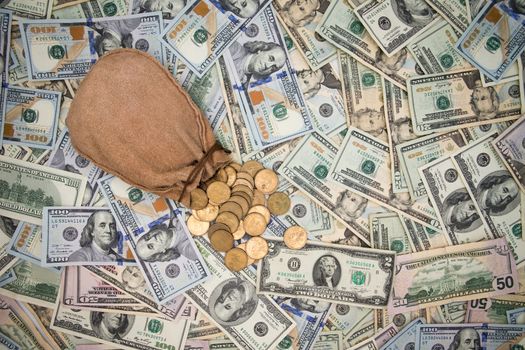 Background of American 100 dollar banknotes scattered randomly completely covering the surface with a hessian bag of spilling dollar coins on top of it, overhead view