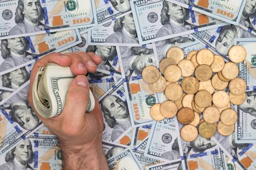 Conceptual financial image of a man clutching a wad of dollar bills over a background of multiple scattered new 100 dollar bills with a pile of dollar coins, closeup overhead view