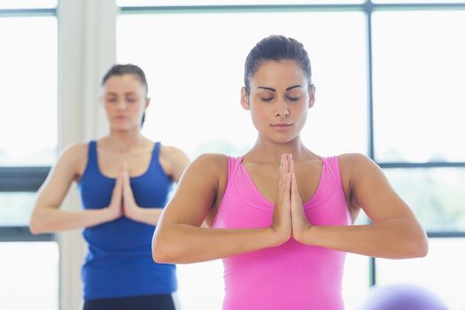 Two sporty young women in Namaste position with eyes closed at a bright fitness studio