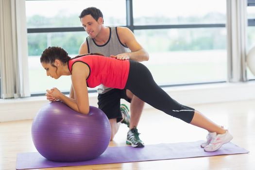 Side view of a trainer helping young woman with fitness ball at a bright gym