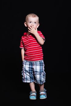 Little boy in a striped shirt and checkered shorts standing with hand in his pocket and hand in mouth