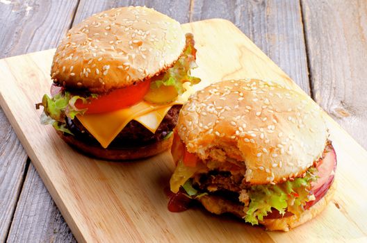 Two Ripe Hamburgers with Beef, Tomato, Lettuce, Pickle, Red Onion and Cheese into Sesame Buns Full Body and Nipped closeup on Wooden Cutting Board