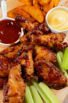 Juicy Chicken Legs and Wings Barbecue with French Fries, Ketchup, Cheese Sauce and Celery Sticks closeup on Wooden Board. Top View