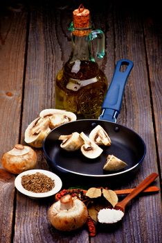 Preparing to Roast Edible Champignon Mushrooms in Frying Pan with Spices in Wooden Spoons, Raw Ingredients and Olive Oil closeup on Rustic Wooden background