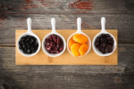 Small ceramic dishes of dried cranberries, apricot, blueberries and cherries arranged in a row on a wooden board on an old weathered wood surface for a healthy snack or tasty appetizer, overhead view