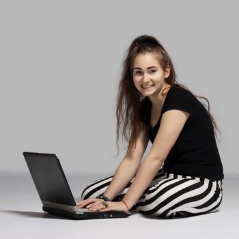Long-haired teenager girl in striped pants sitting in cross-legged on the floor and working on laptop