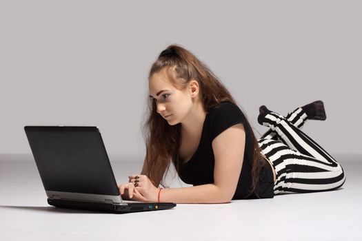 Long-haired teenager girl in striped trousers lying on the floor and working on laptop