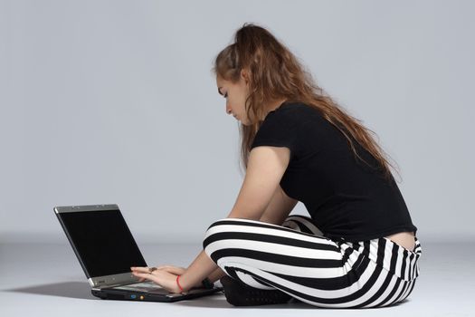 Long-haired teenager girl in striped pants sitting in cross-legged on the floor and working on laptop