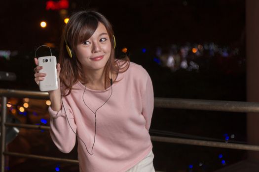 Attractive Malaysian female wearing headphones and holding a cell phone with city lights in background