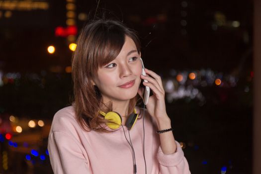 Attractive Malaysian female with headphones and speaking on smart phone with city lights in background
