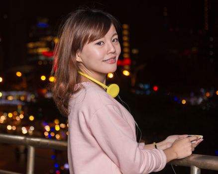 Attractive Malaysian female wearing headphones and holding a cell phone with city lights in background