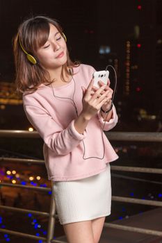 Attractive Malaysian female wearing headphones and holding a cell phone with city lights in background