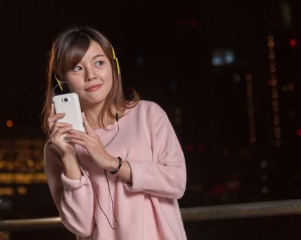 Attractive Malaysian female wearing headphones and holding a cell phone with city lights in background