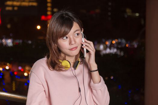 Attractive Malaysian female with headphones and speaking on cell phone with city lights in background