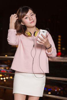 Attractive Malaysian female wearing headphones and holding a cell phone with city lights in background