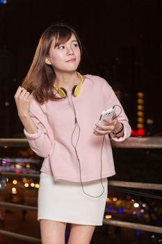 Attractive Malaysian female wearing headphones and holding a cell phone with city lights in background