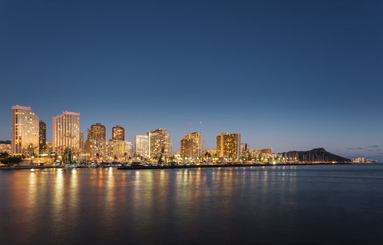 Panorama of the nightime skyline of Honolulu and Waikiki from Ala Moana park after the sun sets with lights illuminating the facades of the hotels and apartments