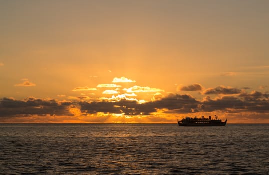 Sunset cruise ship sails on horizon as the sun sets slowly behind clouds off Oahu in Hawaii