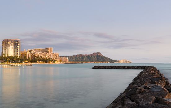 Panorama of the skyline of Waikiki at dusk taken with a long exposure to blur out movement in the water. Breakwater leads the eye towards the volcano of Diamond Head in Hawaii