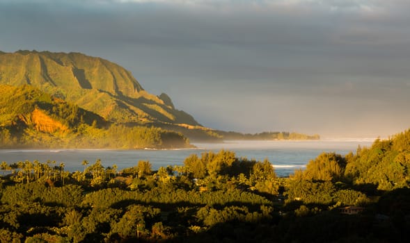 Bay at Hanalei in Kauai with the Na Pali mountain range in the background. Taken just after dawn with the rising sun lighting the peaks of the mountains and casting a sunbeam ray into the ocean