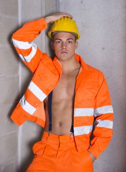Handsome young construction worker with orange suit open on naked torso, wearing yellow hardhat