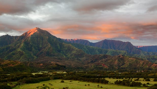 Mountains of the Na Pali mountain range in the distance above Hanalei Bay in Kauai. Taken just after dawn with the sun just skimming the peaks of the mountains and illuminating the clouds with a brilliant red light