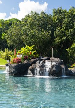 Hot tropical swimming pool at resort with waterfall feature and reclining chairs on the patio with turquiose water