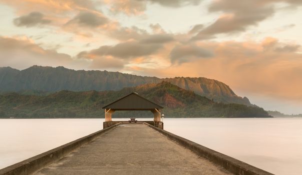 Rising sun illuminates the peaks of Na Pali mountains over the calm bay and Hanalei Pier in long exposure photo